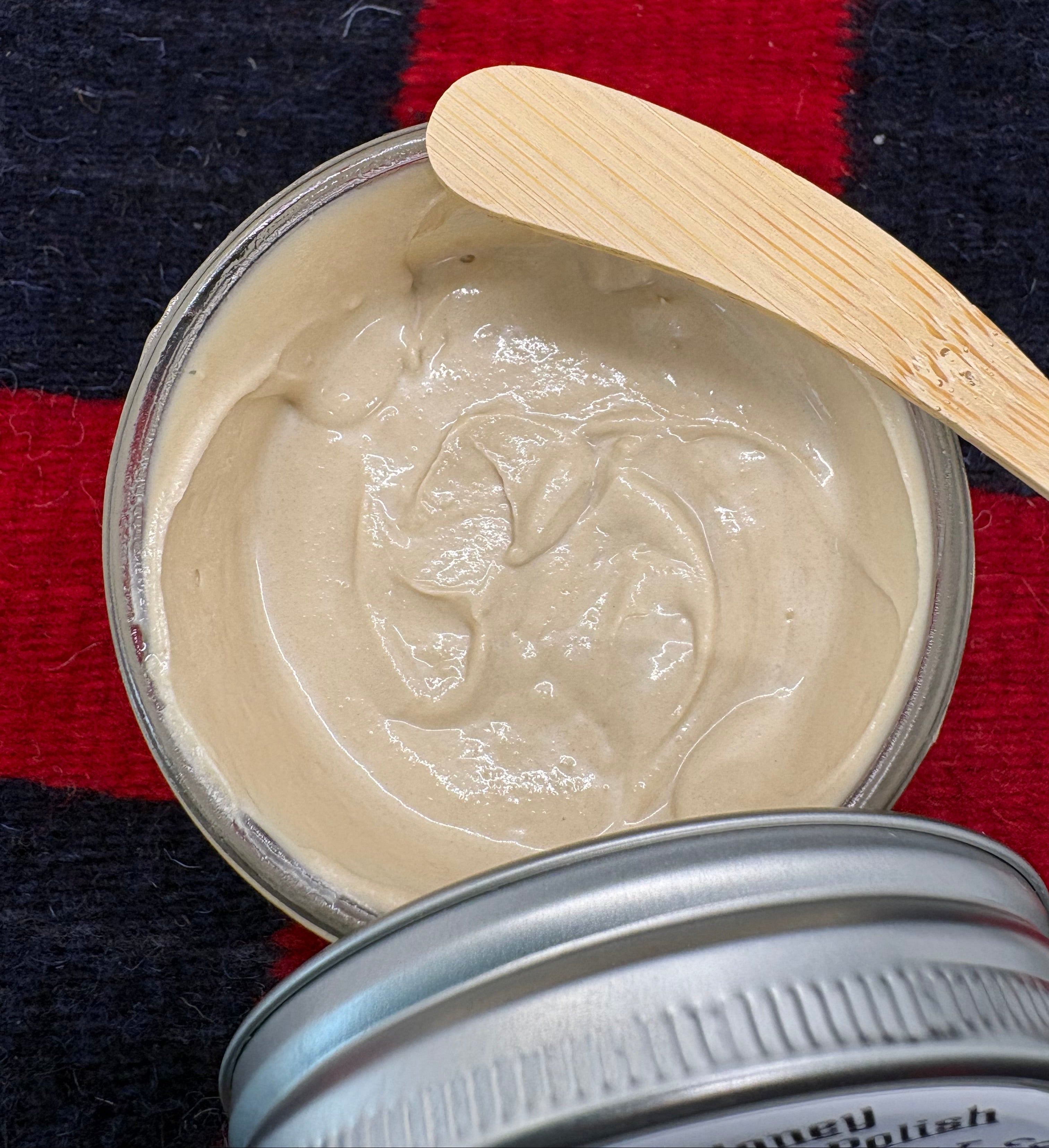 Open jar of beige cream with a wooden applicator on a red and black striped fabric background